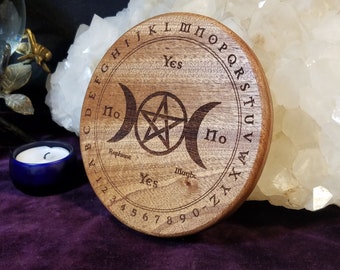 May include: A round wooden divination board with engraved letters, numbers, and symbols. The board features a triple moon design, a pentagram, and the words "Yes," "No," and "Maybe." The board is a light brown color.