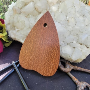 May include: A wooden, teardrop-shaped pendant with a small hole at the top. The pendant has a rich, reddish-brown color and a textured surface. The pendant is likely for jewelry or craft projects.