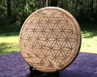 May include: A circular wooden decorative item featuring an engraved Flower of Life design. The natural wood has dark etched lines. It's displayed on a black stand, set against a backdrop of greenery and a purple tablecloth. The diameter is approximately 30 cm.