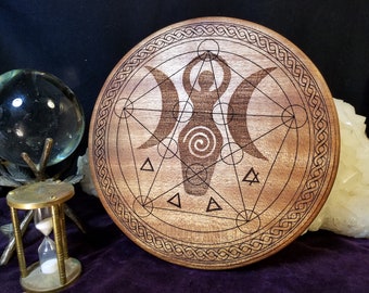 Op de afbeelding: Een ronde houten decoratie met een gesneden ontwerp. Het toont een centrale vrouwelijke figuur, halve manen, geometrische vormen en een rand. Het hout heeft een warme, natuurlijke kleur. Waarschijnlijk voor spirituele of decoratieve doeleinden.
