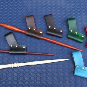 May include: A collection of wooden wands in various colors, including red, brown, and white, paired with leather holsters in black, brown, green, red, and blue. The holsters are secured with gold-colored rivets, set against a blue background.