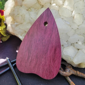 May include: A purpleheart wood pendant with a teardrop shape and a small hole at the top. The wood grain is visible, and the pendant has a smooth finish. The pendant is a craft item.