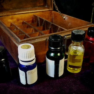 May include: A wooden box with multiple compartments, containing five small glass bottles with different colored liquids. The bottles have black and white caps. The box is open and sitting on a purple surface.