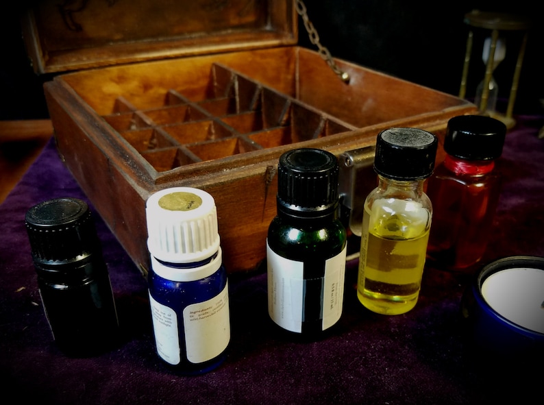 May include: A wooden box with multiple compartments, containing five small glass bottles with different colored liquids. The bottles have black and white caps. The box is open and sitting on a purple surface.