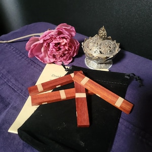 May include: Three wooden sticks with a red and white wood grain pattern. The sticks are arranged in a triangle shape and are sitting on a black velvet pouch. The pouch is on a purple surface. The text "Druid" is visible on a piece of paper in the background.