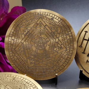 Pode incluir: Disco circular de cor latão envelhecido com padrões geométricos e texto gravados. O design central apresenta uma estrela de cinco pontas dentro de um hexágono. Discos adicionais com designs semelhantes são visíveis.