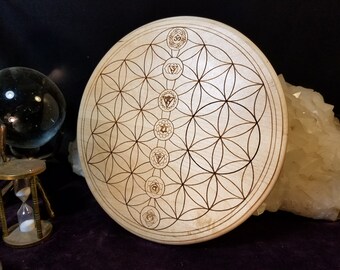 May include: A light-coloured wooden chakra plate featuring a flower of life pattern and chakra symbols. The round plate has a detailed, etched design. The background includes a crystal, a glass ball, and an hourglass.