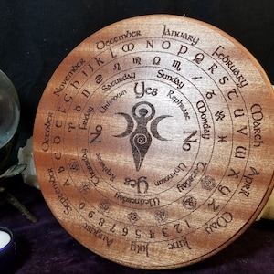 May include: A circular wooden divination board with a triple moon symbol in the centre. The board is engraved with the days of the week, months of the year, and letters of the alphabet.