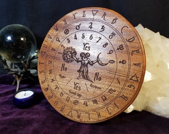 May include: A round wooden divination board with engraved symbols, letters, and numbers. The board features a central illustration of a figure holding a sun and moon. The words "Yes," "No," and "Good-bye" are also engraved on the board.