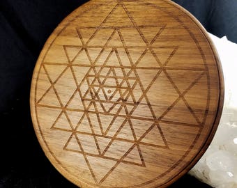 May include: A circular wooden coaster with an etched Sri Yantra design. The coaster is made of light brown wood and features a geometric pattern of interlocking triangles and a central dot. The coaster's diameter is approximately 10 cm.