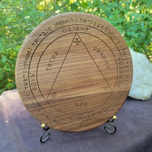 May include: A wooden circular disc with a triangle inscribed inside a circle. The triangle has the words "Tetra", "Gramma", and "NOL" inscribed on its sides. The circle has the words "ORIGNS", "SGYN", "PAYMON", and "AMAYMON" inscribed around the outside. The disc is on a black metal stand.