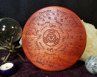 May include: A wooden circular disc with a red-brown stain. The disc has a circular design in the center with a star and a Celtic knot. The outer ring has the months of the year and days of the week written around the edge. The disc is surrounded by a crystal ball, a candle, and a rock.