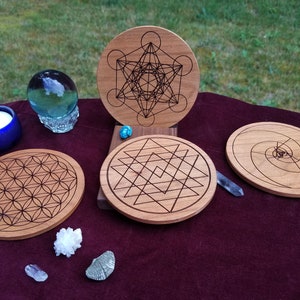 Könnte beinhalten: Holzuntersetzer mit geometrischen Mustern, darunter die Blume des Lebens und Metatrons Würfel. Sie liegen auf einem weinroten Tuch, zusammen mit Kristallen, einem blauen Kerzenhalter und einer Glaskugel. Durchmesser ca. 10 cm.