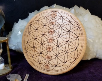 May include: A light-coloured wooden chakra plate featuring a flower of life pattern and seven chakra symbols. The round plate has a detailed, etched design. A large white crystal formation is in the background.