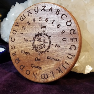 May include: A round wooden Ouija board with engraved letters, numbers, and phrases. The board features a sun design in the center and is surrounded by the alphabet and numbers. The board is made of light brown wood.