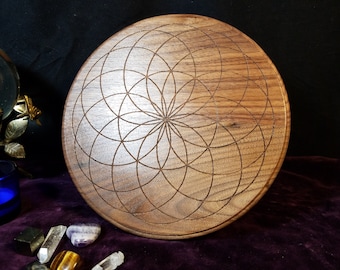May include: A circular wooden tray with an engraved geometric pattern. The tray is made of light brown wood and features a symmetrical design of intersecting circles. The design is centered and covers the entire surface of the tray.