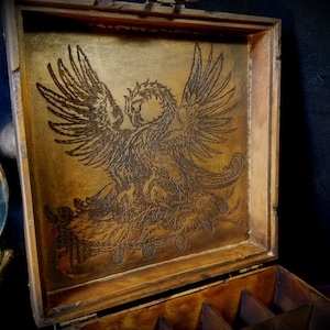 May include: A wooden box with a hinged lid. The inside of the lid is decorated with an intricate laser-etched design of a griffin. The box has multiple compartments inside.