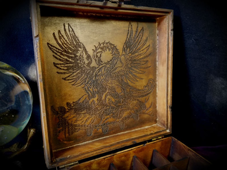 May include: A wooden box with a hinged lid. The inside of the lid is decorated with an intricate laser-etched design of a griffin. The box has multiple compartments inside.