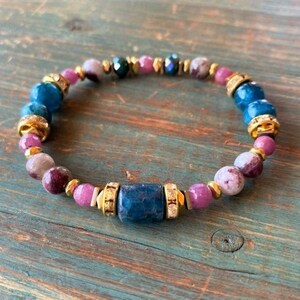 May include: A beaded bracelet featuring a mix of blue, pink, and gold-colored beads. The bracelet includes a central rectangular blue bead, surrounded by round beads in various shades of pink and purple, and gold-colored spacers.