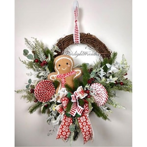 May include: A Christmas wreath with a brown grapevine base, decorated with greenery, red berries, and two large red and white swirl lollipops. A felt gingerbread man and a red and white patterned bow are centered. The text "Starlight Wreaths" is visible.