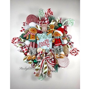 May include: A festive Christmas wreath featuring gingerbread men, lollipops, and ribbons in red, white, and green. A blue sign reads "Christmas Cookie Cafe". The wreath is decorated with various textures and embellishments, creating a cheerful holiday display.