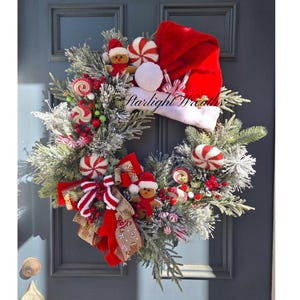 May include: A Christmas wreath featuring flocked pine branches, a red Santa hat, gingerbread figures, red and white peppermint candies, red berries, and a red and white striped ribbon. The text Starlight Wreaths is visible on the hat.