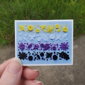 May include: A blue fabric sticker with embroidered yellow, purple, and black floral designs. The sticker is a representation of the non-binary pride flag.