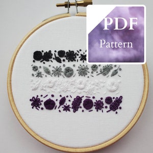 May include: A close-up of a white embroidery hoop with black, gray, and purple embroidery floss. The embroidery depicts a series of floral and leaf designs in a horizontal pattern. The hoop is on a white background with a purple and gray watercolor background with the text "PDF Pattern" in white.