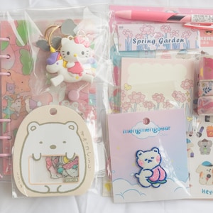 May include: A collection of stationery items in pink and pastel colors. Includes a pink binder, a Hello Kitty keychain, stickers, washi tape, a pen, and a notepad with floral designs. There is also a bear patch and a bear-shaped tag.