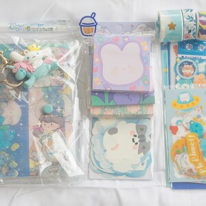 May include: A collection of stationery supplies in a clear plastic binder. Includes stickers, notepads, decorative tape, a pen, and a keychain with a cartoon character. The items have a pastel blue and purple color scheme with cute illustrations.