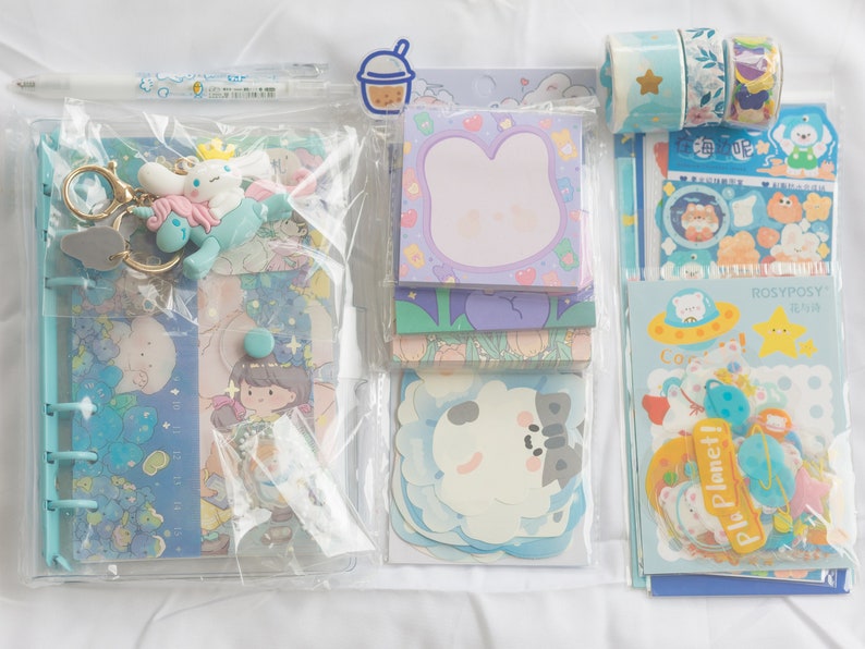 May include: A collection of stationery supplies in a clear plastic binder. Includes stickers, notepads, decorative tape, a pen, and a keychain with a cartoon character. The items have a pastel blue and purple color scheme with cute illustrations.