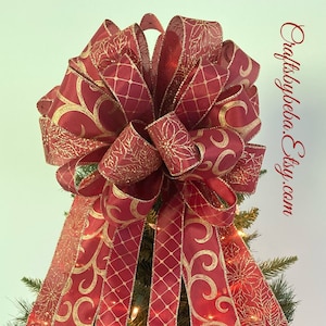 May include: A red and gold Christmas tree topper bow with a swirl pattern and a poinsettia design. The bow is tied with a wired ribbon and has a glittery finish.