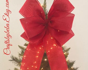 May include: A large, vibrant red bow adorns the top of a Christmas tree, its long ribbons cascading down. The tree is lit with warm white lights, creating a festive holiday display. The Etsy shop name "Craftsbybeba.etsy.com" is visible.