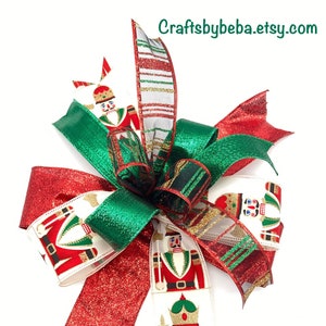 May include: A Christmas bow made with red, green and white ribbon featuring a nutcracker print. The bow is tied with a white ribbon with gold glitter and has a red glitter tail.