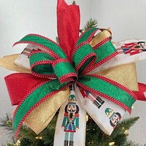 May include: A Christmas tree topper bow with red, gold, and green ribbon. The ribbon is decorated with white polka dots. The bow is accented with a white ribbon with a repeating pattern of nutcracker figures. The ribbon also has a gold glitter trim. The text "Craftsbybelba.etsy.com" is visible at the bottom of the image.