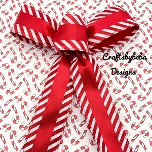 May include: A red and white striped ribbon bow with a glittery finish. The bow is tied on a white background with a repeating pattern of red and white candy canes. The text "Craftsbybeba Designs" is visible in the bottom right corner of the image.