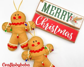 May include: Two felt gingerbread men ornaments with gold string lights wrapped around them. The ornaments are hanging from a string. The background is white with a wooden sign that says "Merry Christmas" in green and red letters.