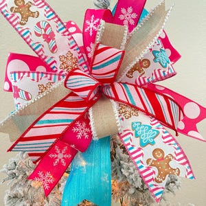 May include: A festive Christmas tree bow featuring a variety of ribbons in bright pink, red, and turquoise. The ribbons are adorned with gingerbread men, candy canes, and snowflakes. The bow is a decorative accent for the holiday season.