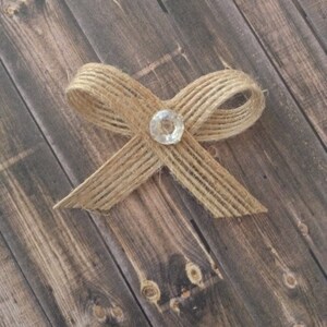 May include: A burlap bow with a clear rhinestone in the center. The bow is on a wooden background.