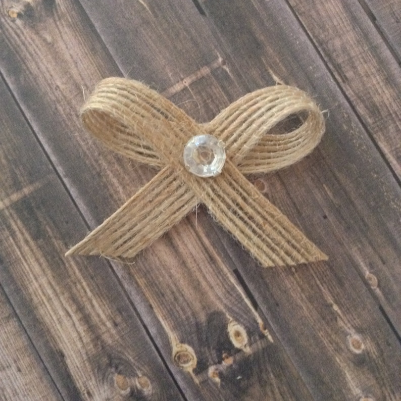 May include: A burlap bow with a clear rhinestone in the center. The bow is on a wooden background.