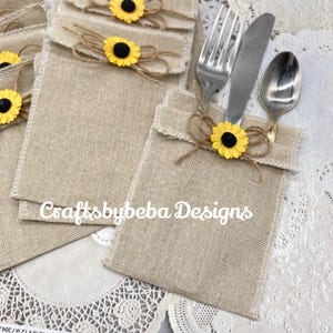 May include: Burlap cutlery holders with a sunflower embellishment and twine bow. The holders are beige with a white stitched border. The text "Craftsbybeba Designs" is printed on the front.