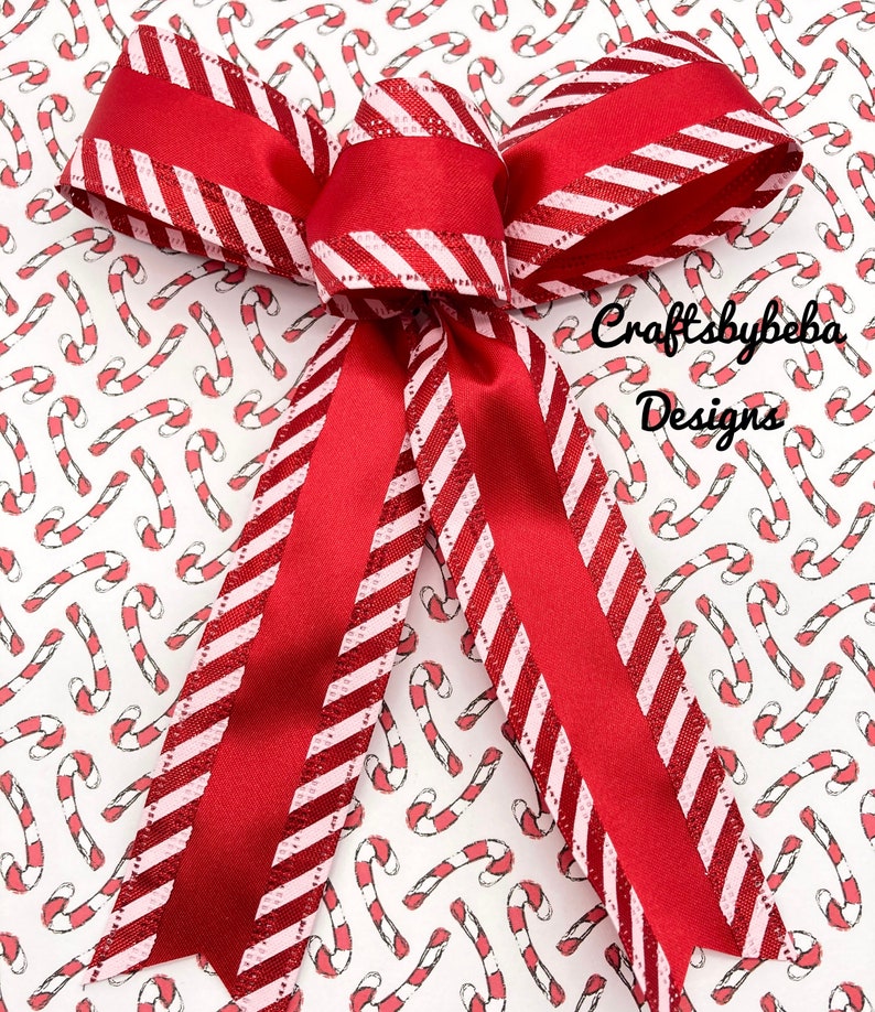 May include: A red and white striped ribbon bow with a glittery finish. The bow is tied on a white background with a repeating pattern of red and white candy canes. The text "Craftsbybeba Designs" is visible in the bottom right corner of the image.
