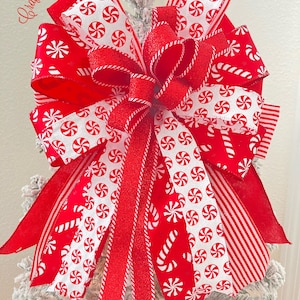 May include: A festive Christmas tree bow in red and white. The bow features multiple layers of ribbon with candy cane and peppermint swirl patterns. The bow is topped with a glittery red ribbon and the text "Craftsbybeba Designs".