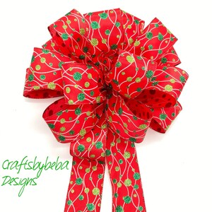 May include: A red and green Christmas bow with a glittery finish. The bow features a pattern of green and gold circles connected by wavy lines. The bow is tied with a long tail.