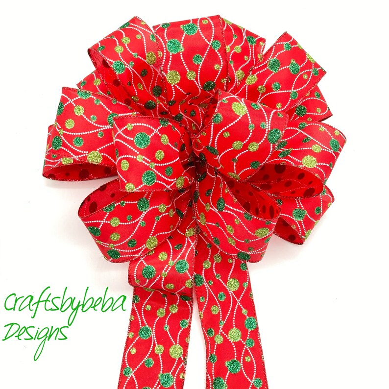 May include: A red and green Christmas bow with a glittery finish. The bow features a pattern of green and gold circles connected by wavy lines. The bow is tied with a long tail.