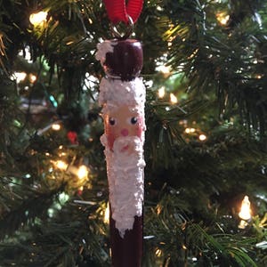May include: A hand-painted wooden Santa Claus ornament with a white beard and a red hat. The ornament is hanging from a red ribbon on a Christmas tree.