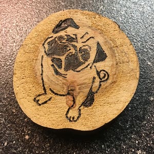 May include: A wood-burned image of a pug dog on a round wooden slice.