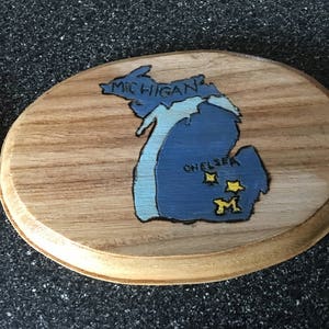 May include: A wooden oval-shaped sign with a blue and white painted outline of the state of Michigan. The sign says "MICHIGAN" and "CHELSEA" with three yellow stars and a yellow "M" inside the state outline.