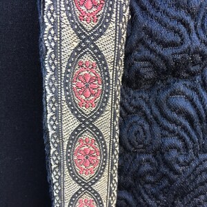 May include: A black fabric with a decorative trim featuring a repeating pattern of oval shapes with pink floral designs. The trim is a combination of black, white, and silver threads.