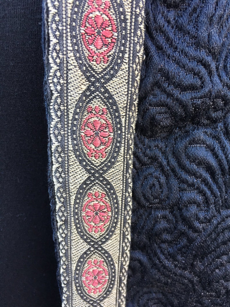 May include: A black fabric with a decorative trim featuring a repeating pattern of oval shapes with pink floral designs. The trim is a combination of black, white, and silver threads.